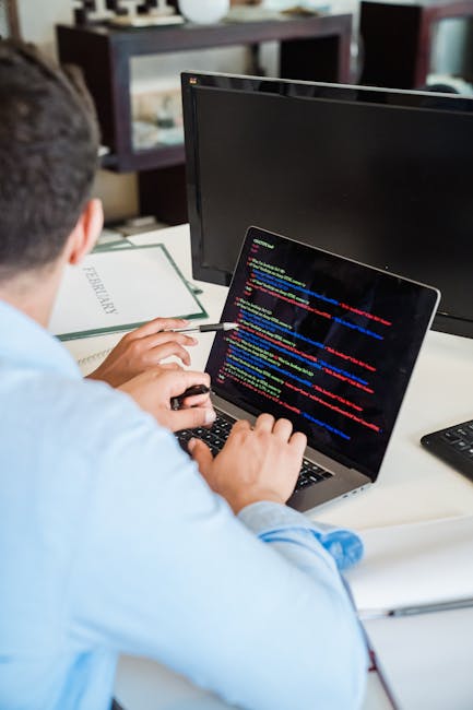 A person wearing a light blue shirt is seated at a desk, working on a laptop displaying a screen filled with colorful lines of code. The workspace features a black monitor to the right of the laptop,