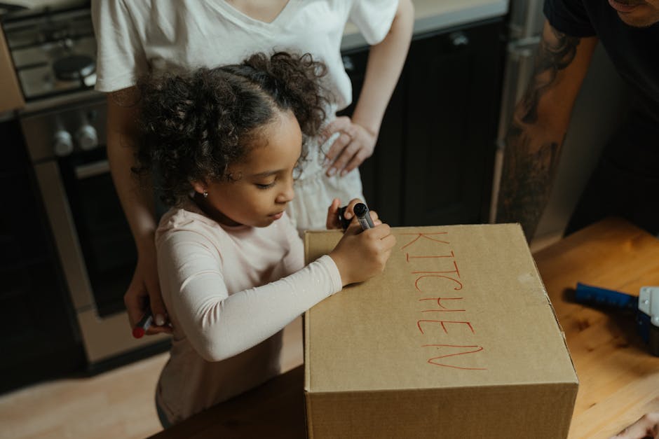 A person in a beige long-sleeved shirt is carefully wrapping a glass plate with transparent bubble wrap on a wooden kitchen countertop, preparing fragile items for a house move. To the left, there is