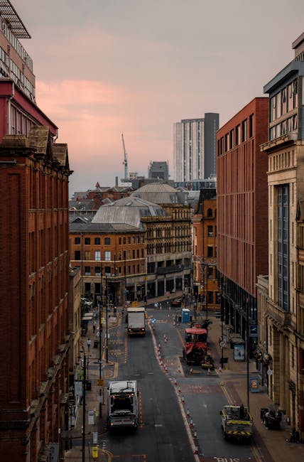 Photograph of a busy urban street scene during daytime in a city centre, featuring a mix of historic and modern buildings with detailed facades, large glass windows, and varying architecture styles. T