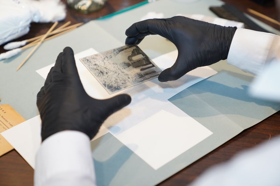 A person wearing black gloves and a white long-sleeved shirt carefully handling a small, flat, rectangular piece of artwork or antique photograph, placing it on a white sheet of paper atop a light blu
